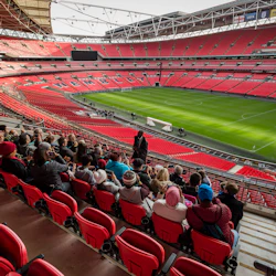 En grupp människor sitter i röda stadionsäten med utsikt över en tom fotbollsplan i en stor, modern stadion.