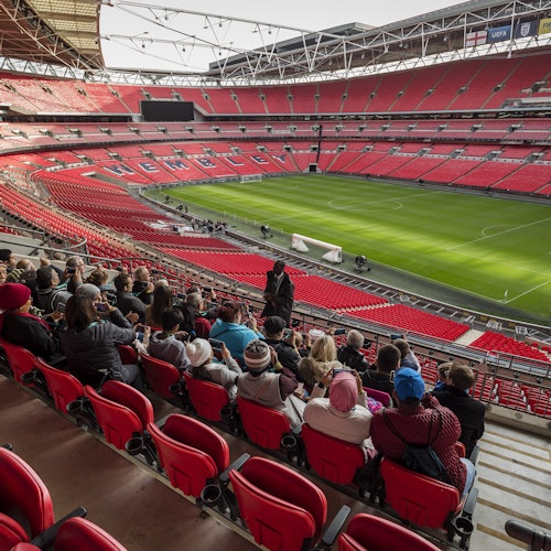 Eine Gruppe von Menschen sitzt auf roten Stadionsitzen mit Blick auf ein leeres Fuu00dfballfeld in einem grou00dfen, modernen Stadion.