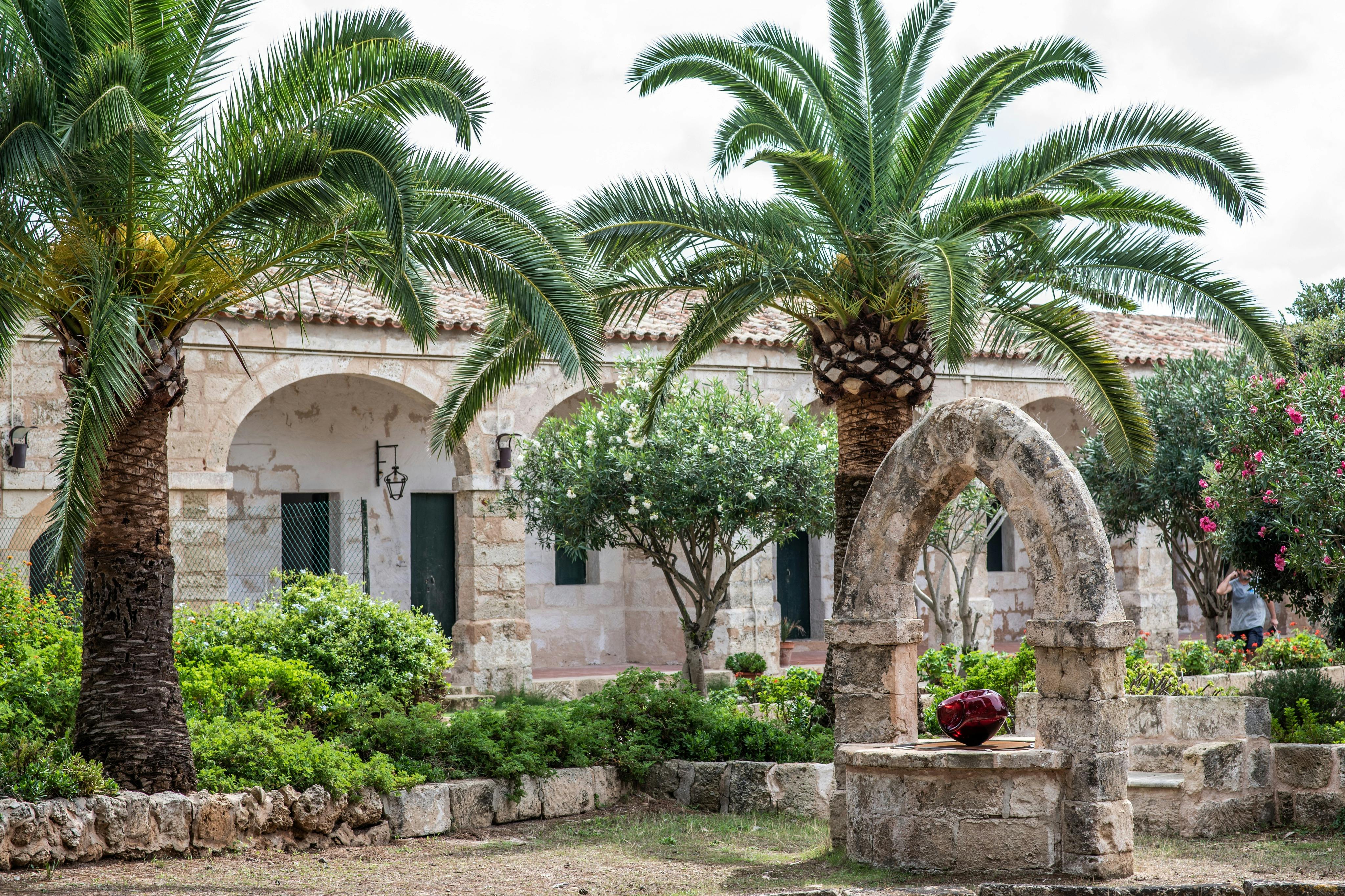 Steinhof mit Palmen, einem gewölbten Brunnen und einer roten Keramikvase. Im Hintergrund ist ein altes Gebäude mit gewölbten Türöffnungen zu sehen.