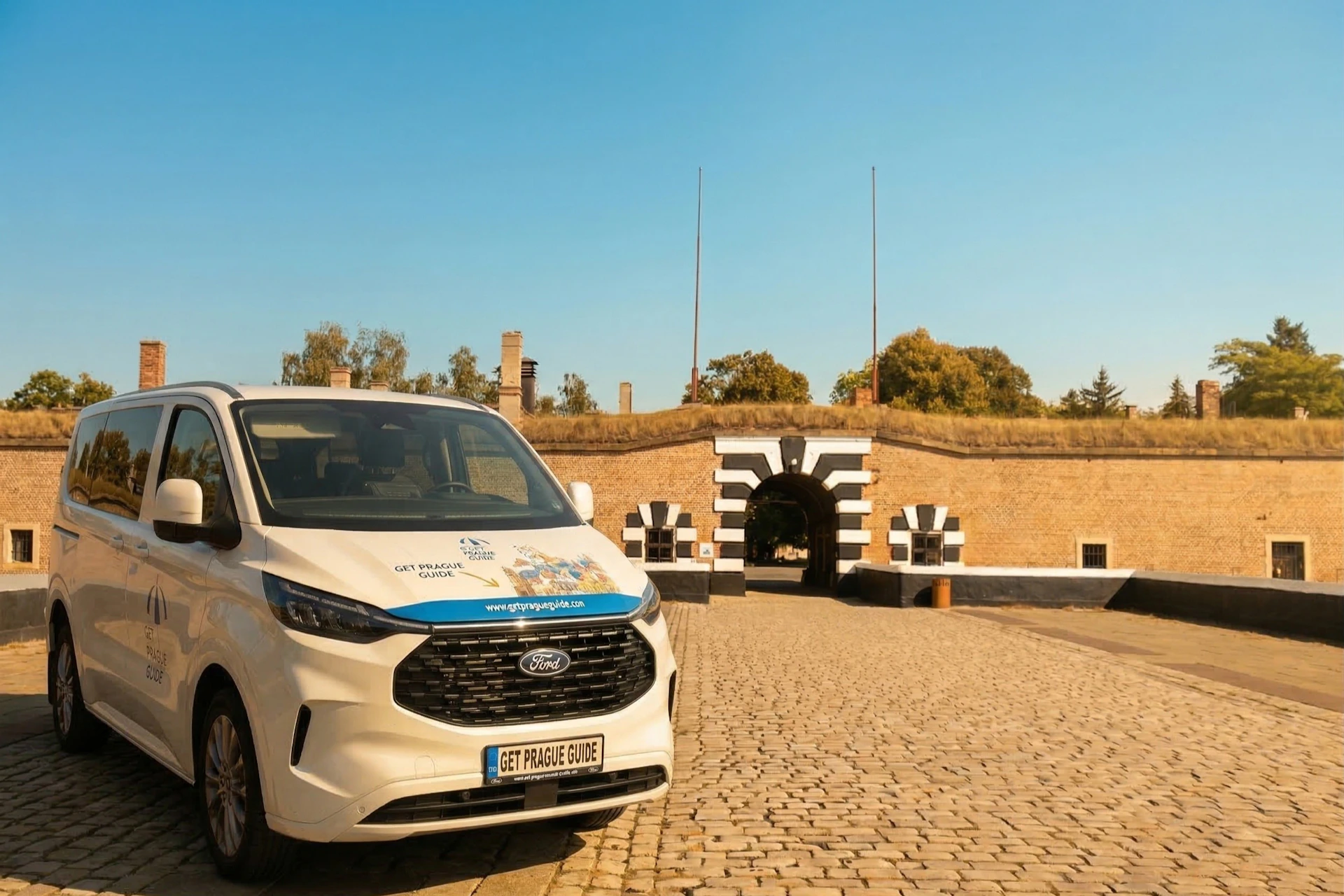 A white van parked on a cobblestone path near a historic fort entrance with striped decoration and flags on poles.