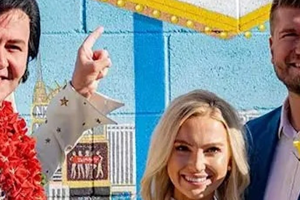 Three people standing in front of a mural with "NEVADA" written on it. The man on the left wears an Elvis costume. The couple beside him hold flowers.