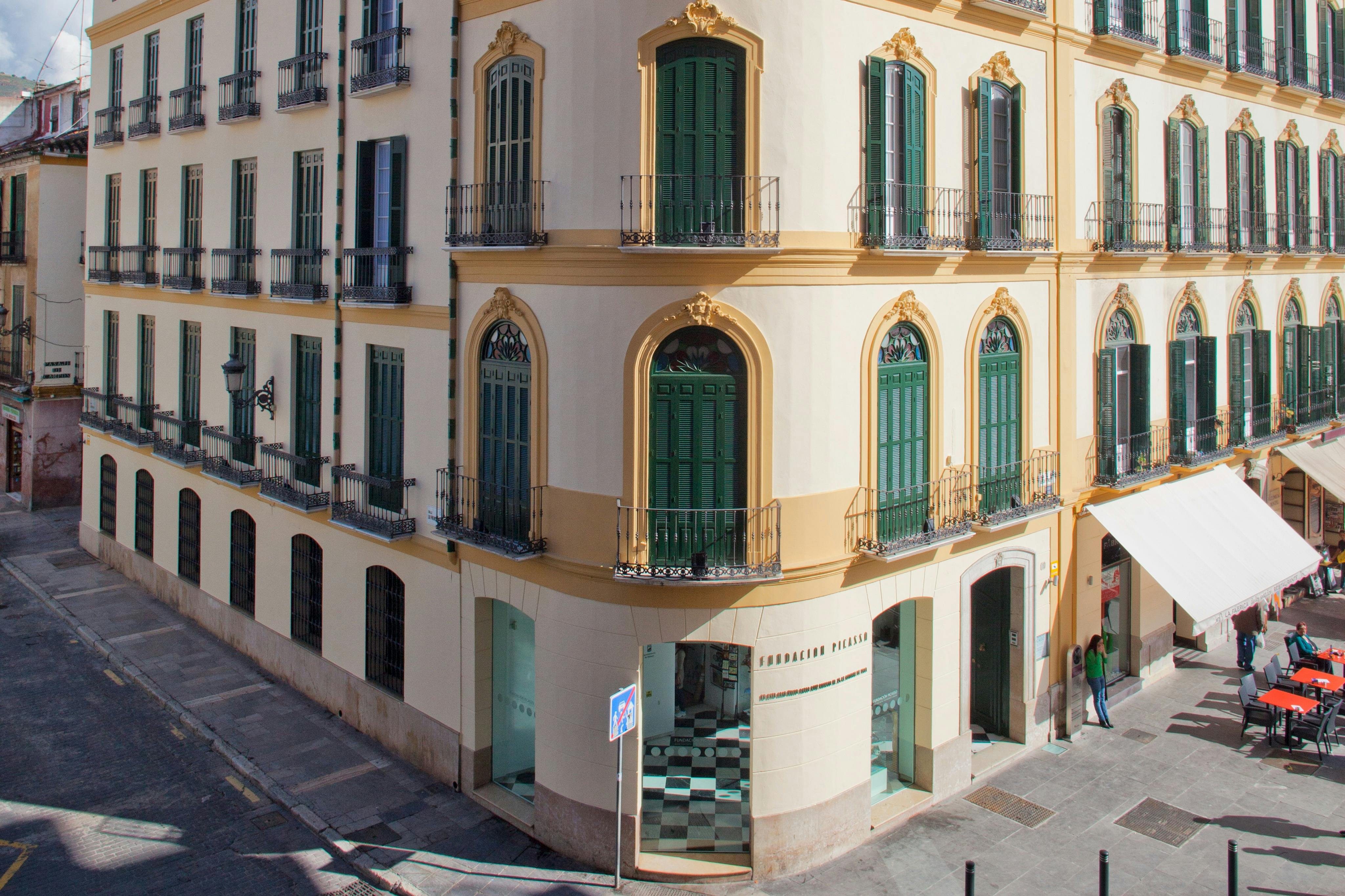 Γωνία ενός μπεζ κτιρίου με πράσινα παράθυρα, τοξωτές πόρτες στο επίπεδο του δρόμου και περίτεχνες αρχιτεκτονικές λεπτομέρειες.