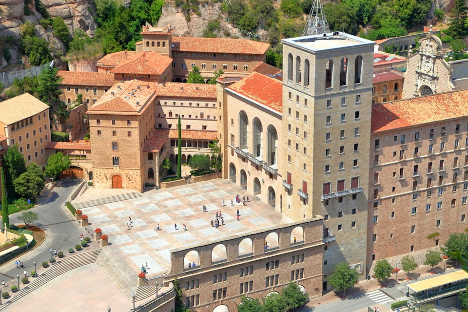 Aerial view of a large historic building complex with a square, arched windows and red-tiled roofs, and people walking in the courtyard.