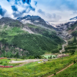 Ett rött tåg färdas genom frodiga gröna berg med ett slingrande spår och en dal med byggnader inbäddade under snötäckta toppar.