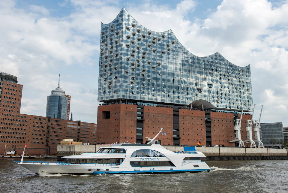 A modern boat sails past the Elbphilharmonie Hamburg, a large, wave-shaped glass building with a brick base, under a partly cloudy sky.