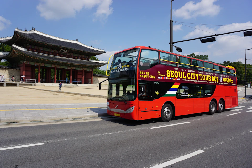 A red double-decker Seoul City Tour Bus passing by a traditional Korean building under a clear blue sky.