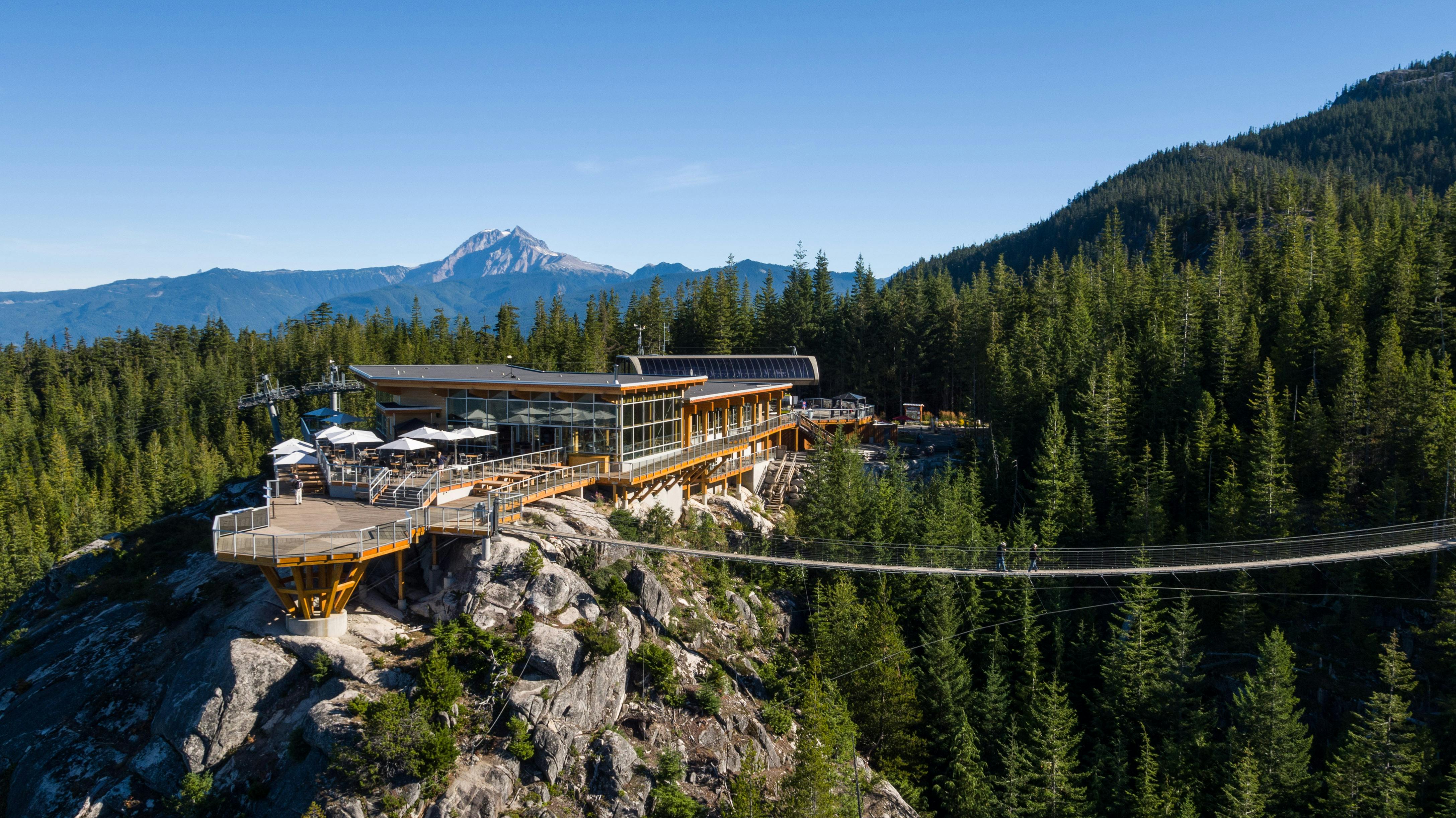 A modern lookout building on a mountain ridge with outdoor seating, surrounded by dense forest and a suspension bridge.