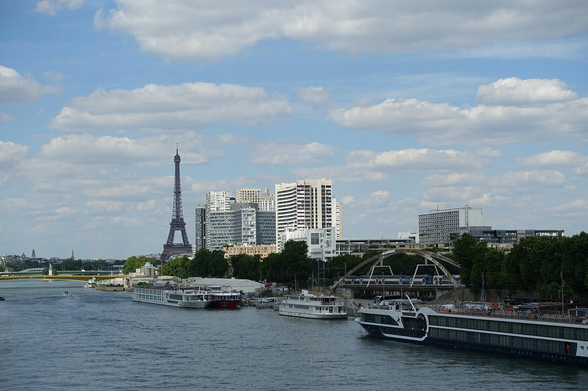 The Eiffel Tower with modern buildings and boats docked along the Seine River under a partly cloudy sky.
