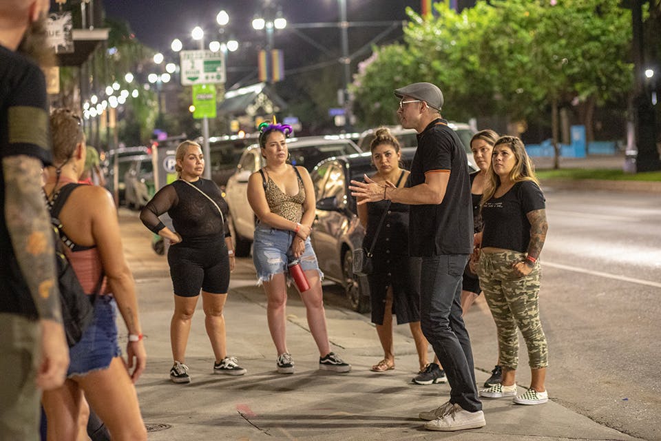 A man speaks to a group of seven women on a city sidewalk at night. They are standing near parked cars under streetlights.