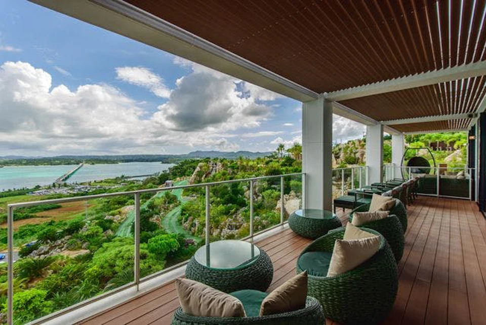 Terrace with green wicker chairs and beige cushions, overlooking lush greenery, rocks, and a body of water under a cloudy sky.