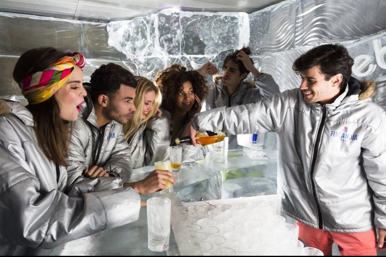 Un groupe de personnes vêtues de vestes argentées boit un verre dans un bar dont les murs et le comptoir sont ornés de décorations en glace.