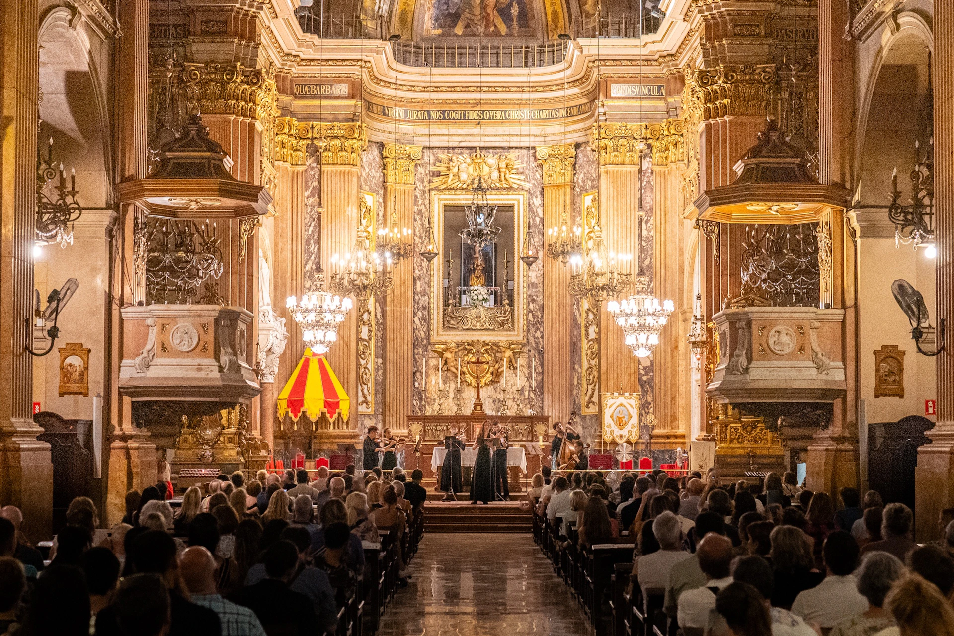 A view of an ornate church interior with chandeliers, golden decorations, and a congregation seated during a ceremony.