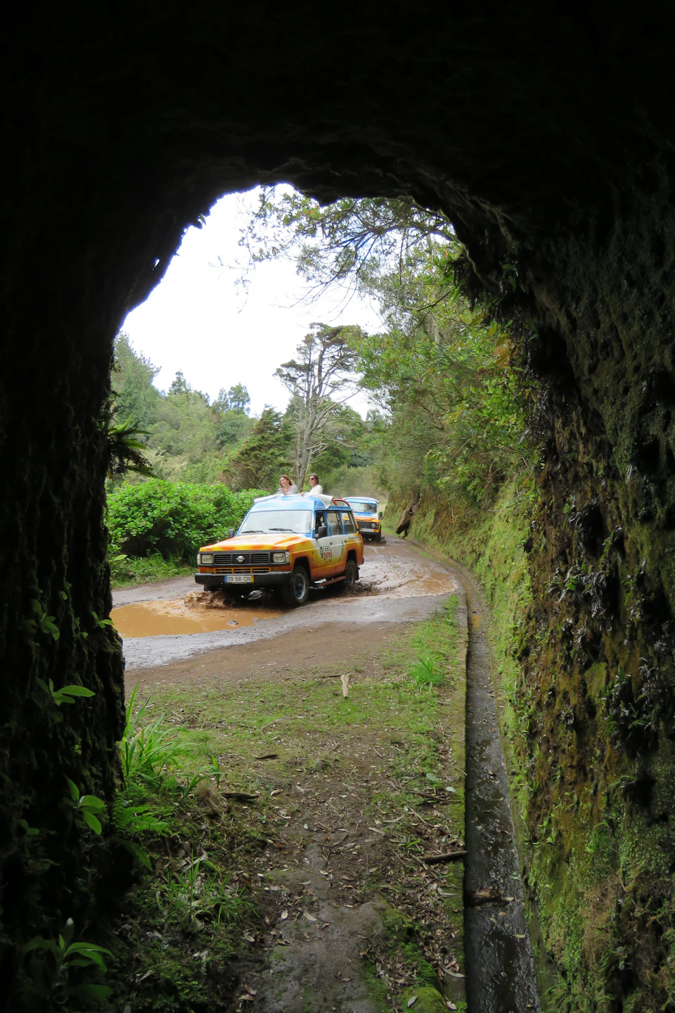 Two off-road vehicles driving through a muddy path, seen from inside a tunnel framed by greenery.