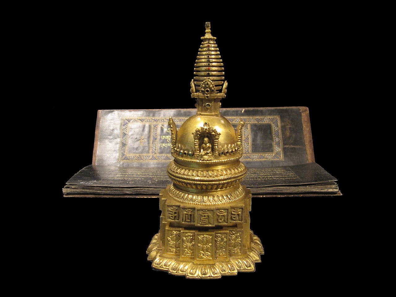 A golden stupa with intricate details is positioned in front of an open, ornate book, set against a black background.
