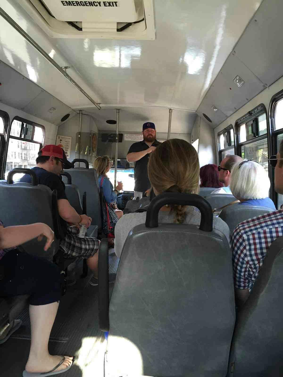 A man is standing and speaking to seated passengers inside a bus. An emergency exit sign is visible on the ceiling.