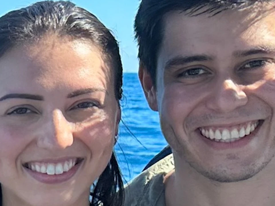 A couple smiling on a boat with a clear blue ocean and a distant city and mountain in the background under a sunny sky.