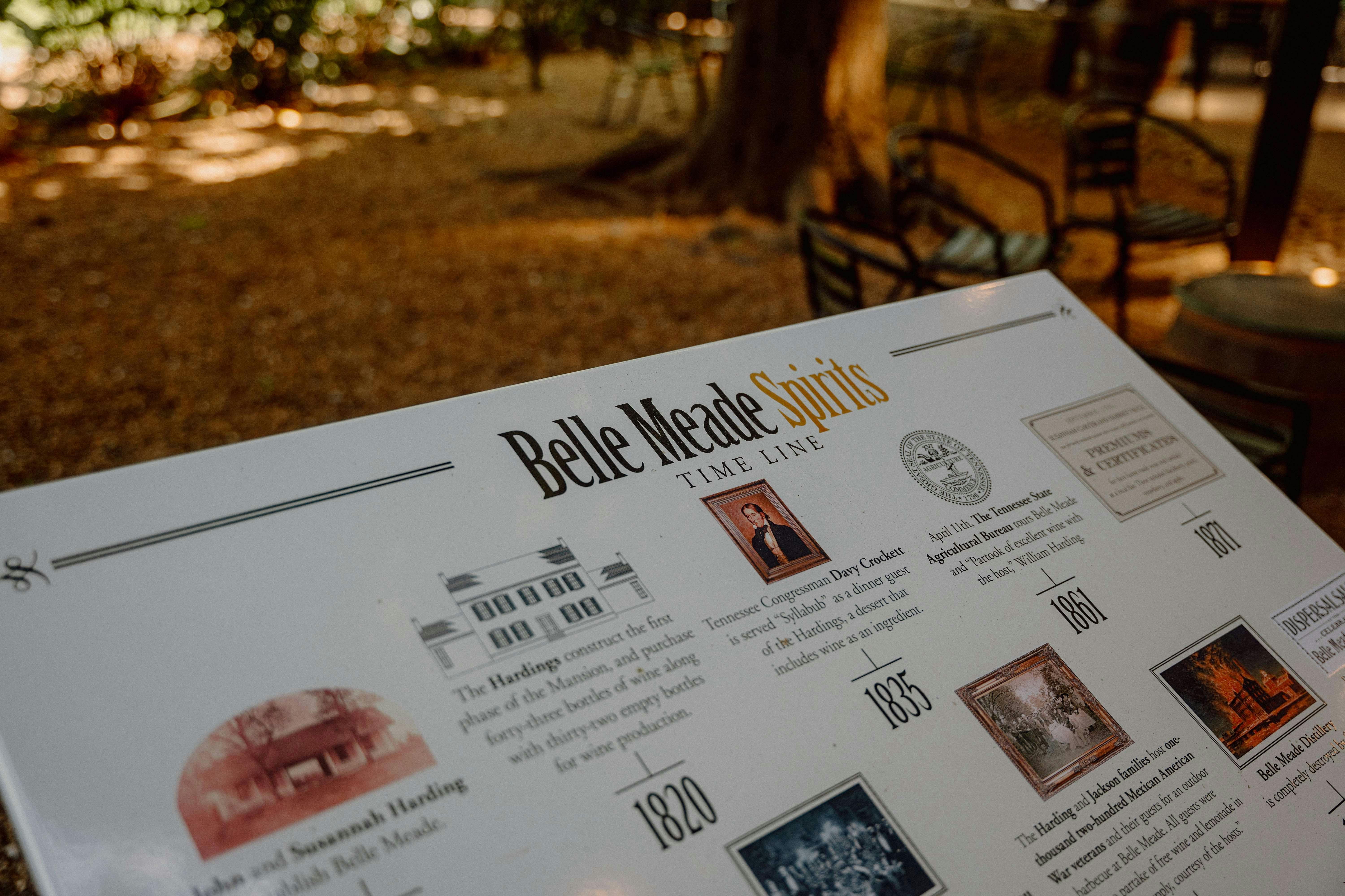 Plakát s časovou osou s názvem „Belle Meade Spirits“ s podrobnostmi o historických událostech s rozmazaným venkovním posezením v pozadí.