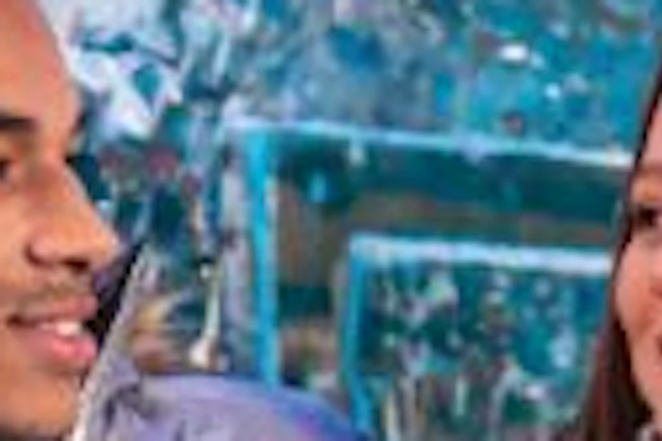 Two people wearing silver puffer jackets sit on an ice bench in an ice bar, one holding a drink and smiling at the other.
