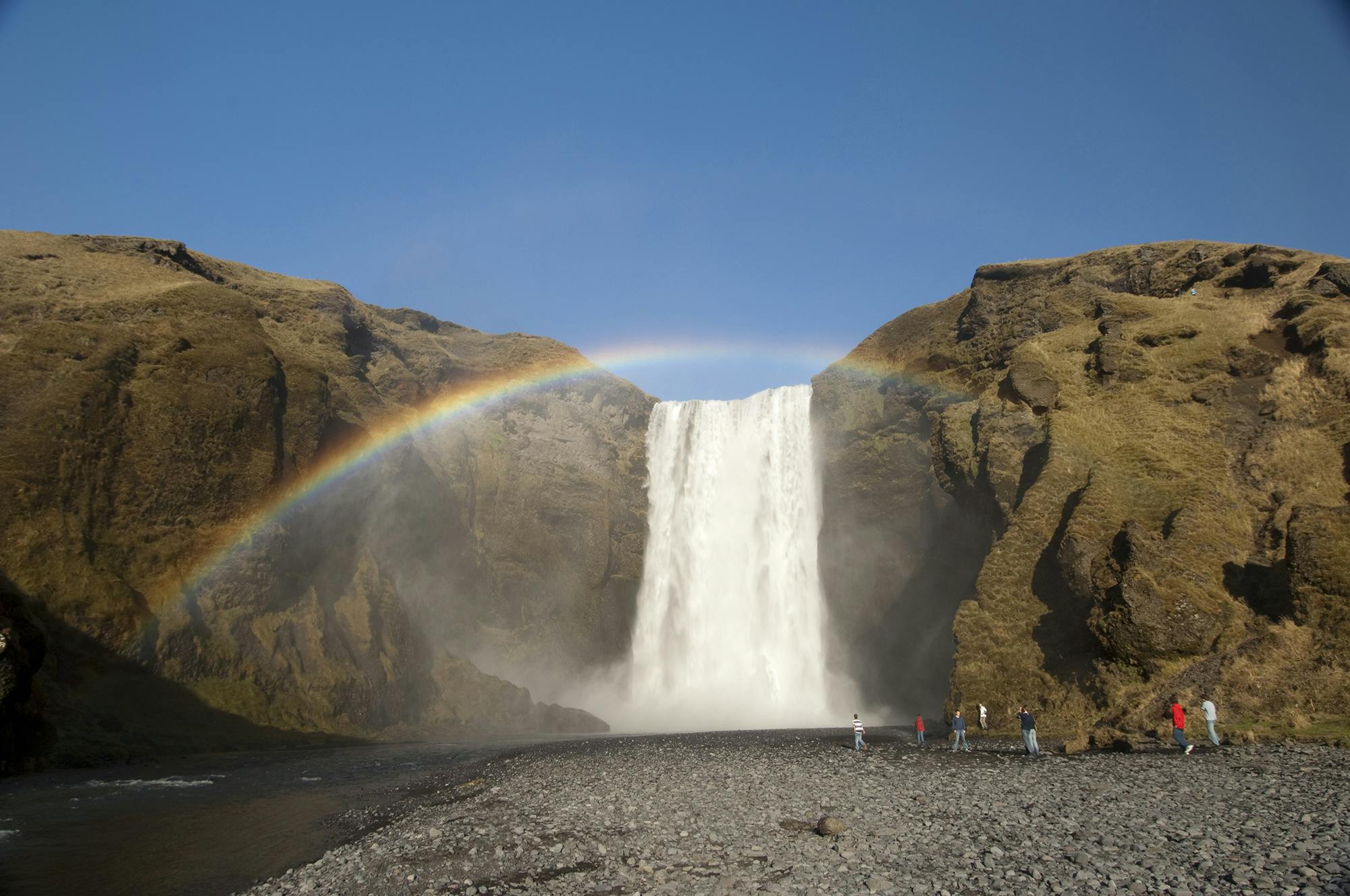 A waterfall cascades between rocky cliffs under a clear sky with a rainbow arching above, and people stand at the base on a rocky ground.