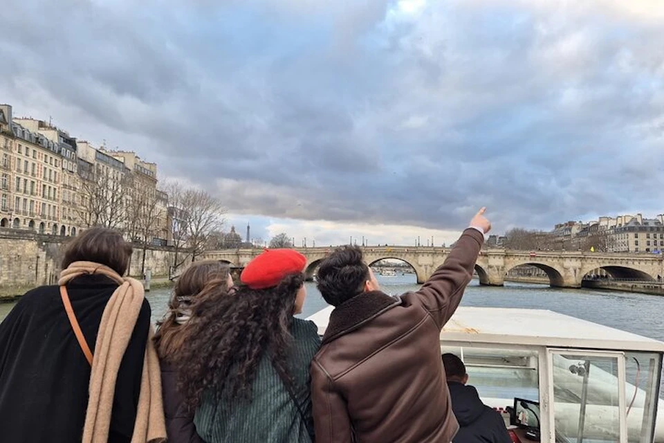 A group of people on a boat points towards a stone bridge under a cloudy sky, with buildings visible in the background.