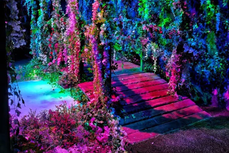 Деревянный мост, украшенный яркими разноцветными цветами и листвой, освещенный яркими огнями, создает сюрреалистическую садовую сцену.