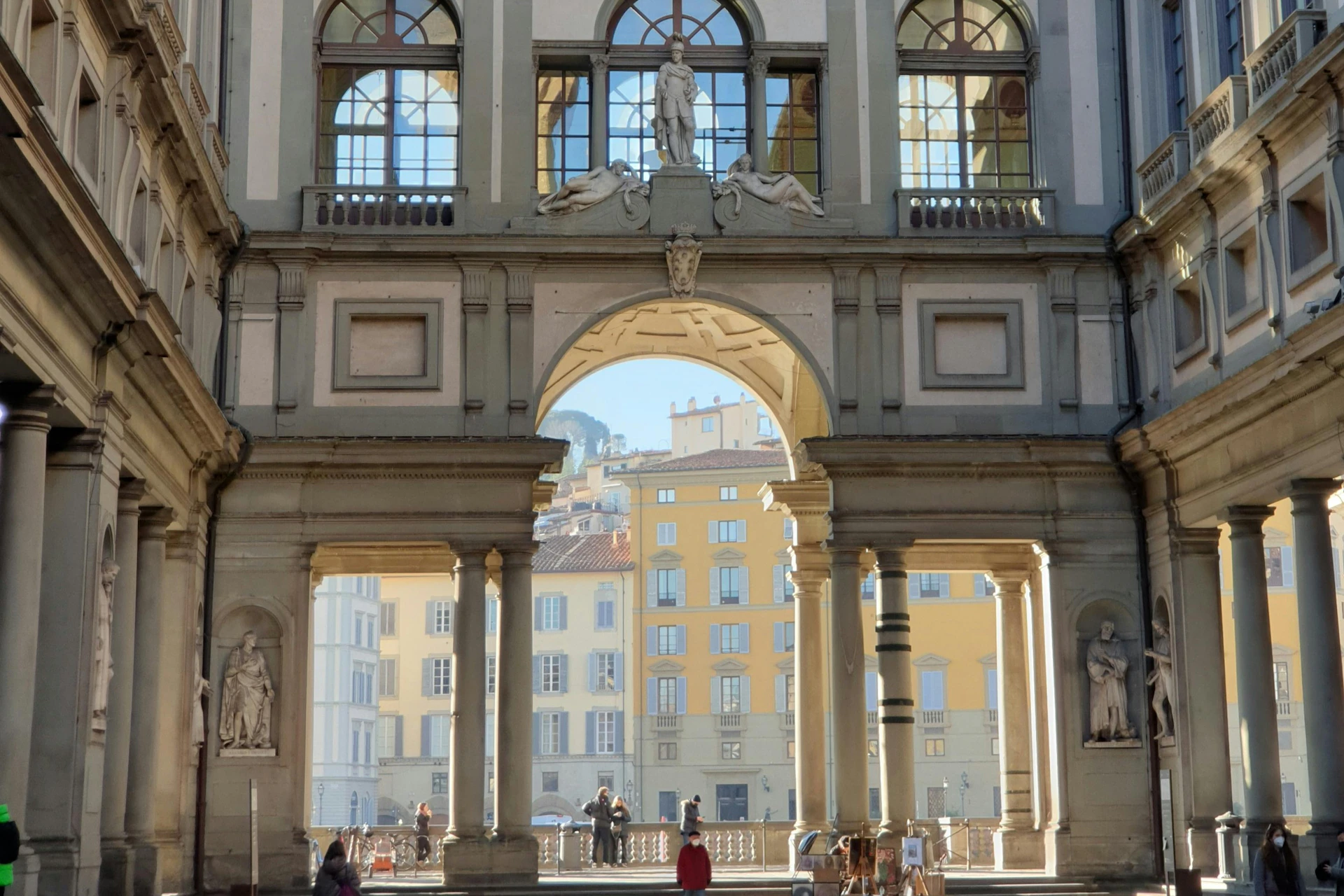 Ornate archway framing a view of yellow buildings in the distance, with statues and people walking underneath.