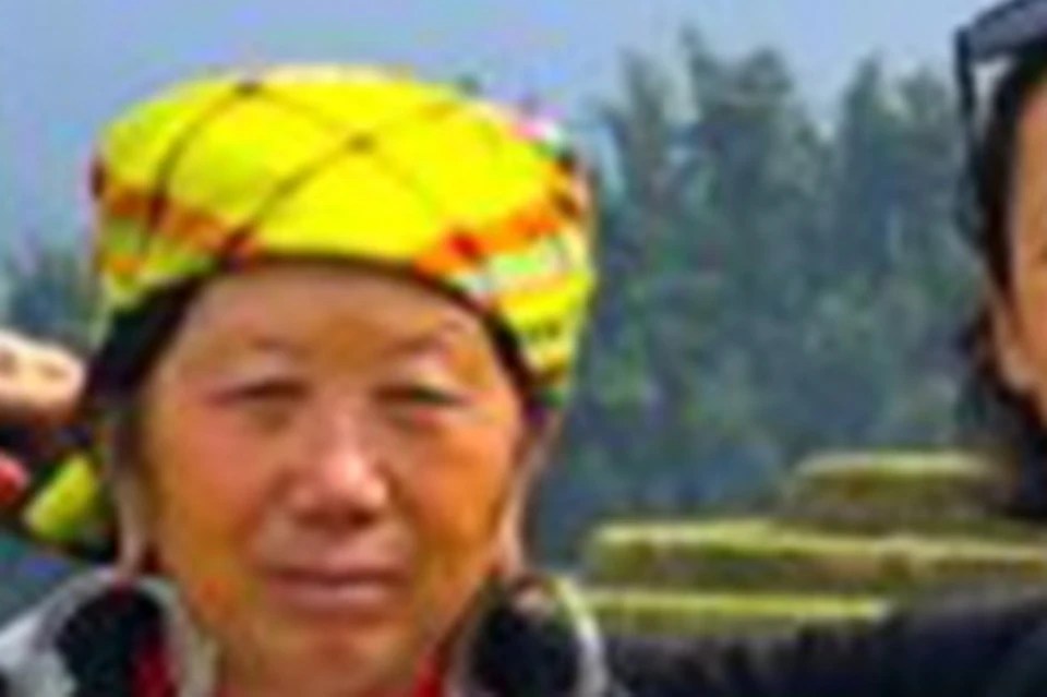 Four women stand in front of terraced fields on a mountainside, with cloudy skies and lush, green hills in the background.