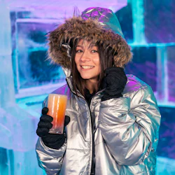 En person i en silverfärgad jacka och handskar ler medan han håller en drink och står framför en isstruktur med färgstark belysning.