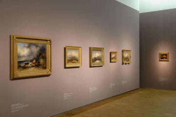 A gallery wall with five framed paintings of various sizes displayed in a row, under soft lighting.
