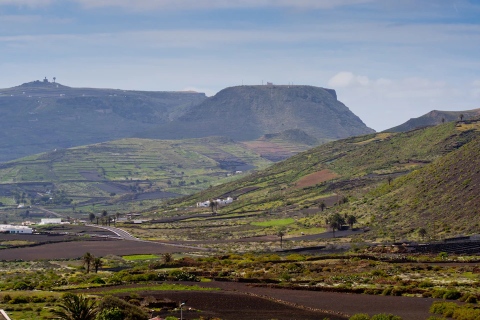 A lush green valley with terraced fields, scattered houses, and a flat-topped mountain under a partly cloudy sky.