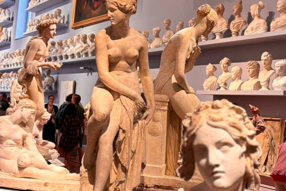 A museum gallery filled with classical sculptures and busts on display, with visitors observing the exhibits.