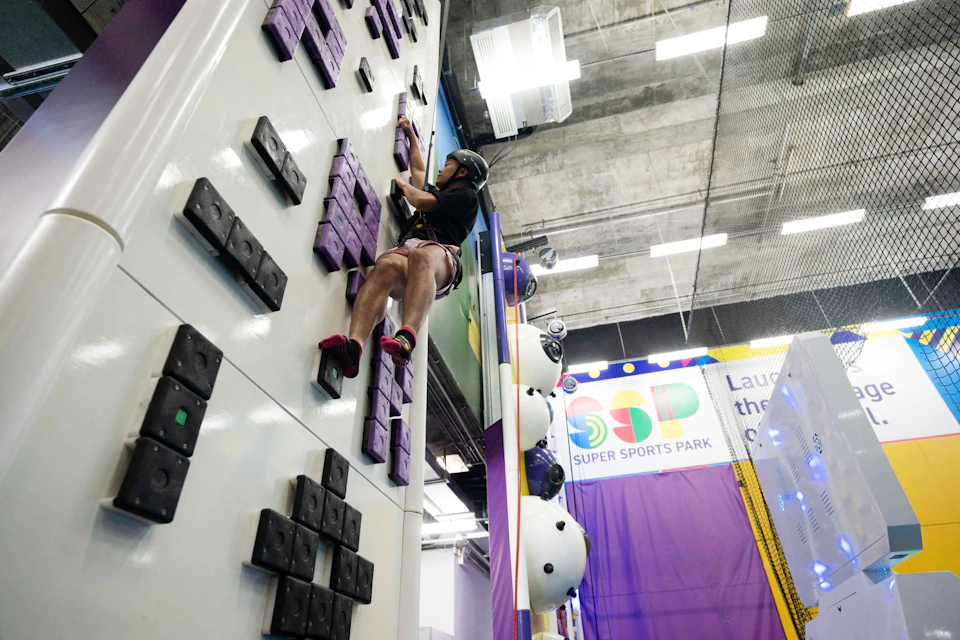 A person wearing a helmet climbs an indoor climbing wall with grips. The background shows a sign for Super Sports Park.