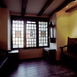 Svagt upplyst rum med trägolv, träbänkar och ett stort fönster med dekorativa glasmålningar.