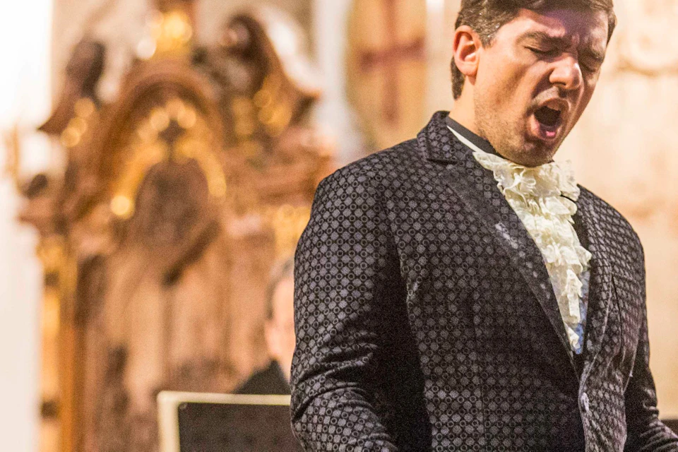 A man passionately singing on stage with musicians playing string instruments in an ornate, classical interior.