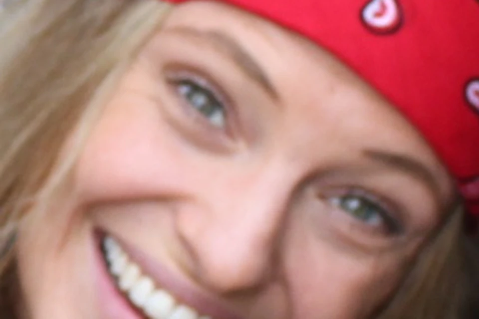 A person wearing a red bandana and grey top crouches and smiles inside a narrow, rocky trench surrounded by dirt and small plants.