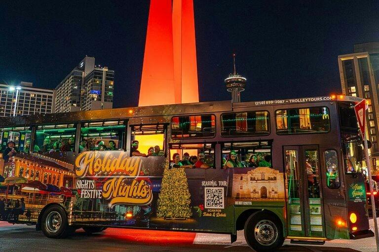 A decorated trolley bus at night with passengers inside and holiday-themed lights, near a tall illuminated tower.
