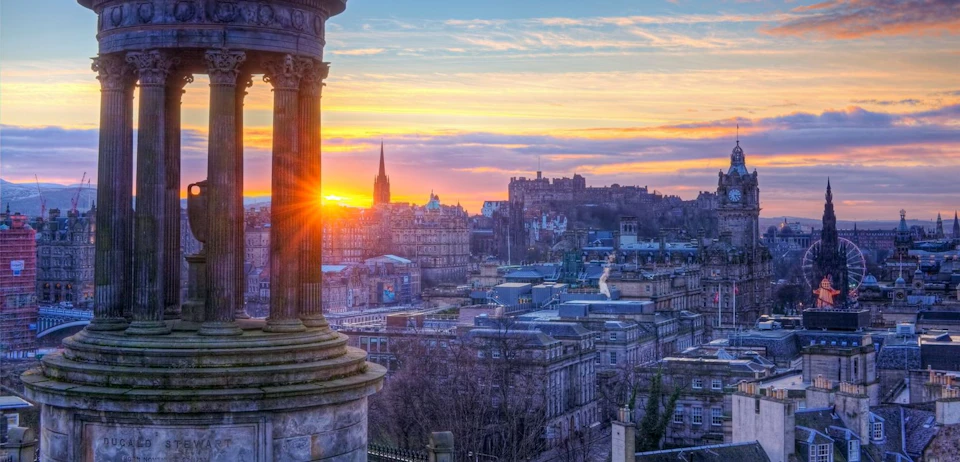 View of an urban skyline at sunset, showcasing historic buildings and a tall spire, framed by a stone column on the left.