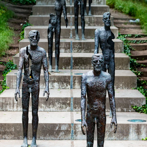 Bronsstatyer som visar mänskliga figurer i olika tillstånd av upplösning, arrangerade på konkreta steg omgivna av grönska.