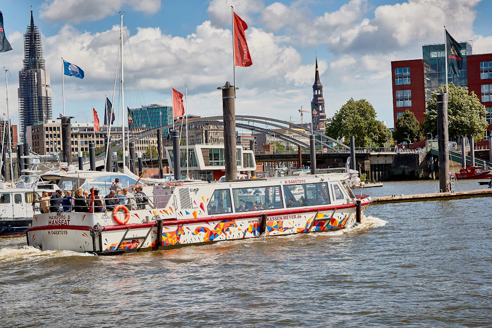 A colorful tour boat with passengers navigates a river, with a bridge, city buildings, and a church tower in the background.
