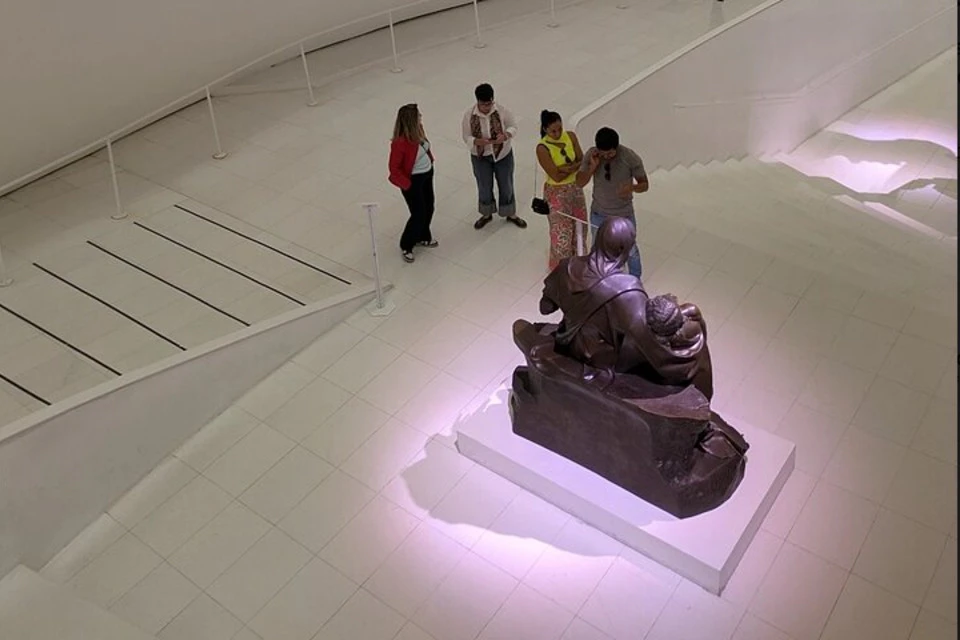 Four people observe a bronze statue in a modern, white-tiled museum with a curved ramp and railing.
