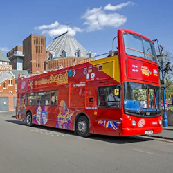 A bright red double-decker tour bus with "City Sightseeing Stratford" graphics, parked on a street near architectural buildings.