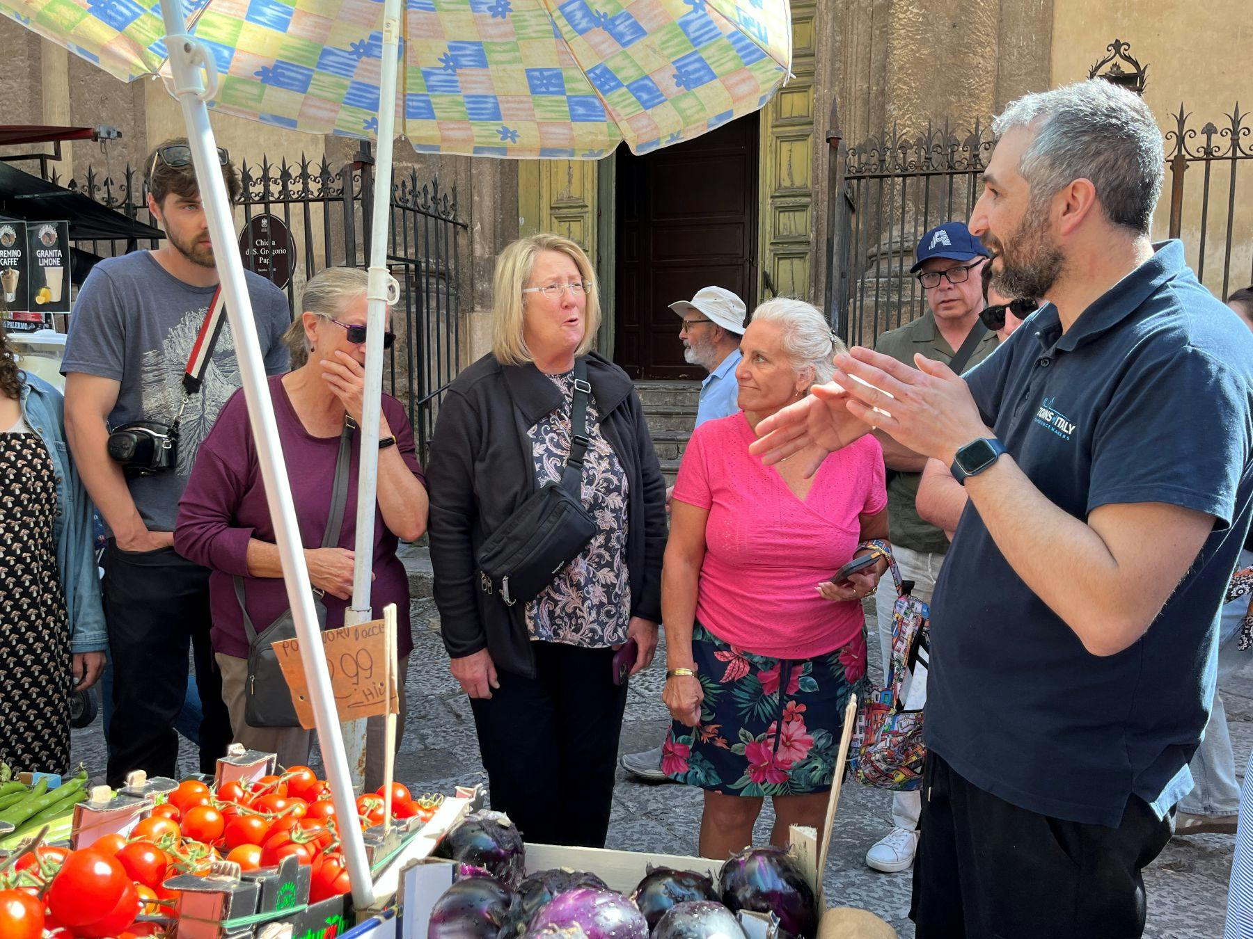 A group of people standing around a market stall with tomatoes and eggplants under a colorful umbrella.