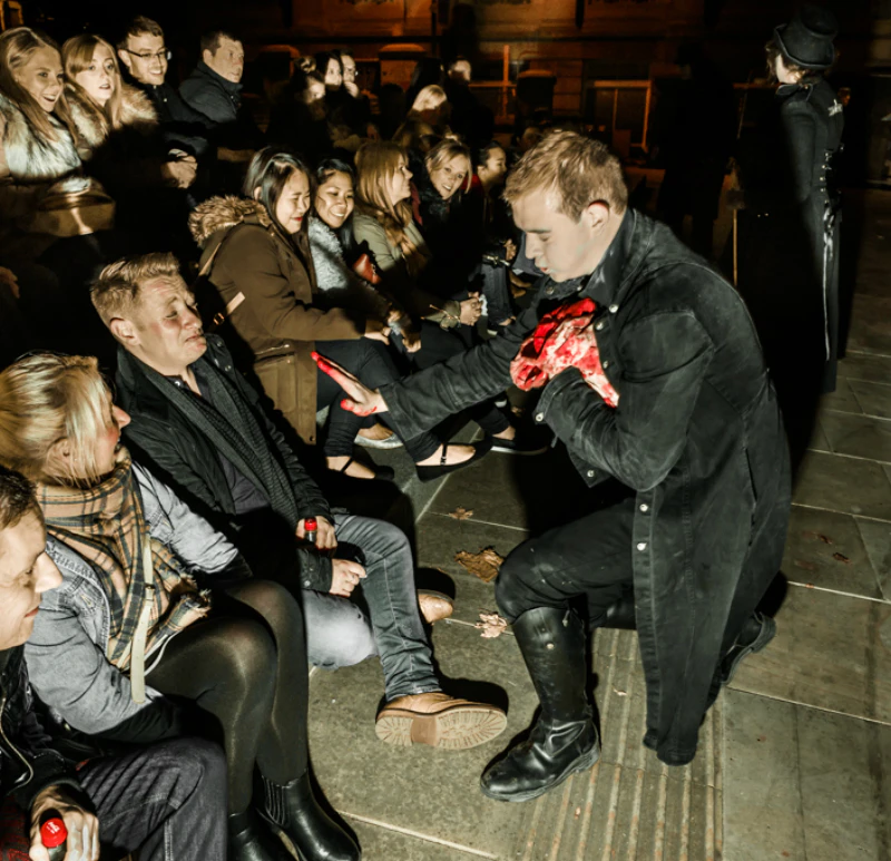 A man in a dark coat performs for a seated audience, holding what appears to be fake organs, while another person stands in the background.