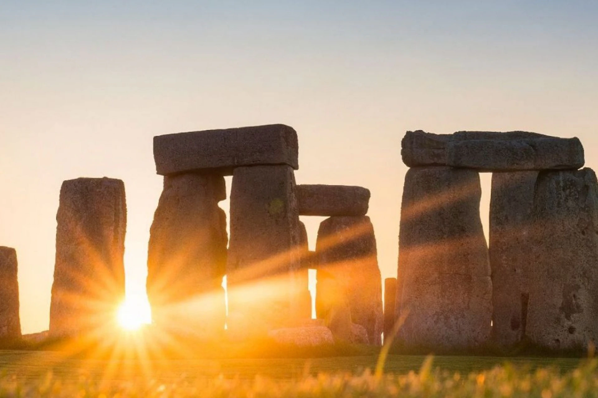Sunlight shining through the stone structures of Stonehenge at sunset, with grass in the foreground.