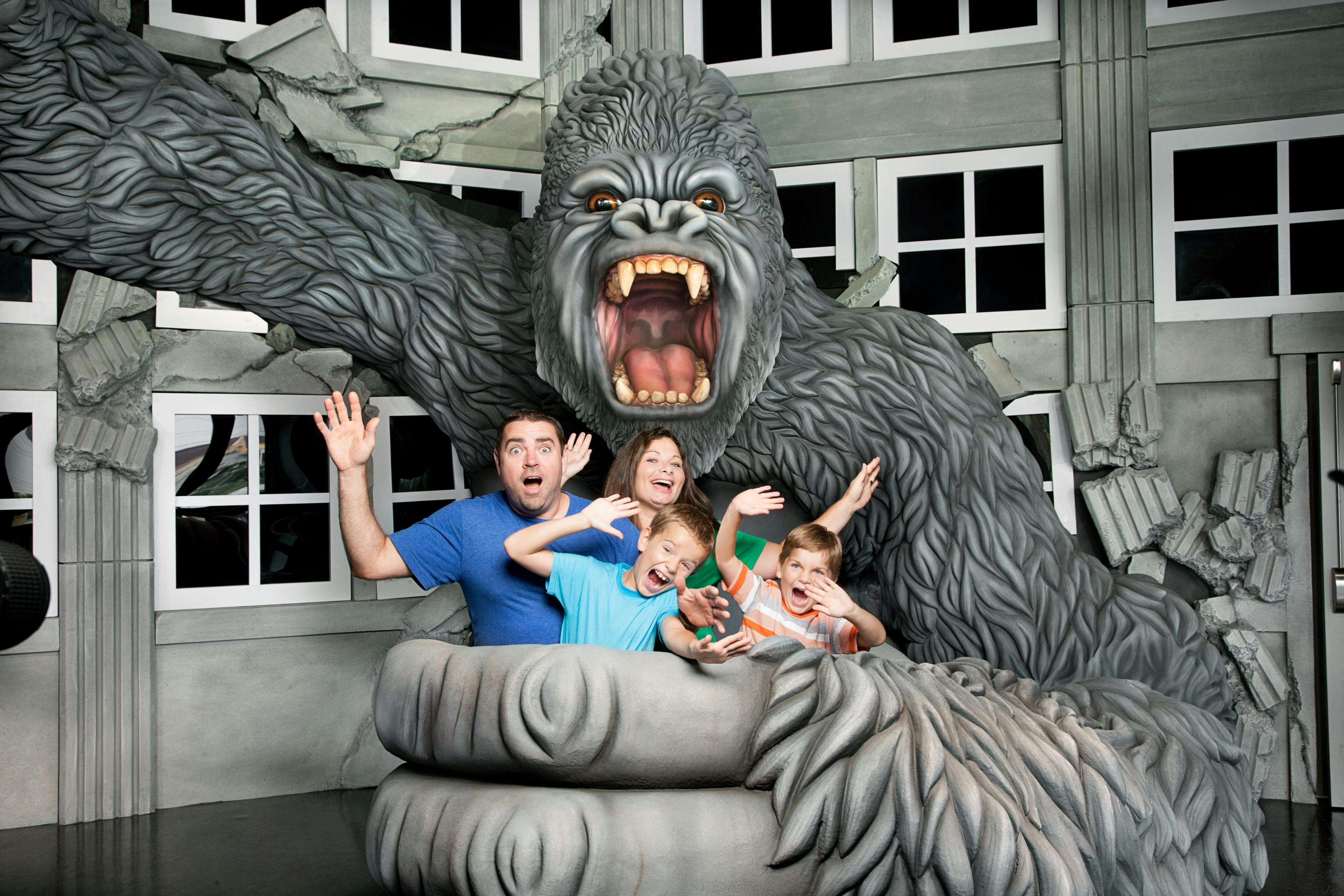Una famiglia di quattro persone posa con un'espressione di sorpresa all'interno di una mano gigante di King Kong sullo sfondo di un edificio.