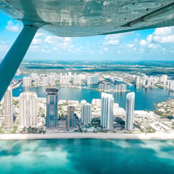 View from an airplane over a coastal city with tall buildings, water bodies, and a beach under a partly cloudy sky.