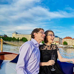 Ett leende par med glasögon njuter av en båttur en solig dag, med byggnader och vatten i bakgrunden.