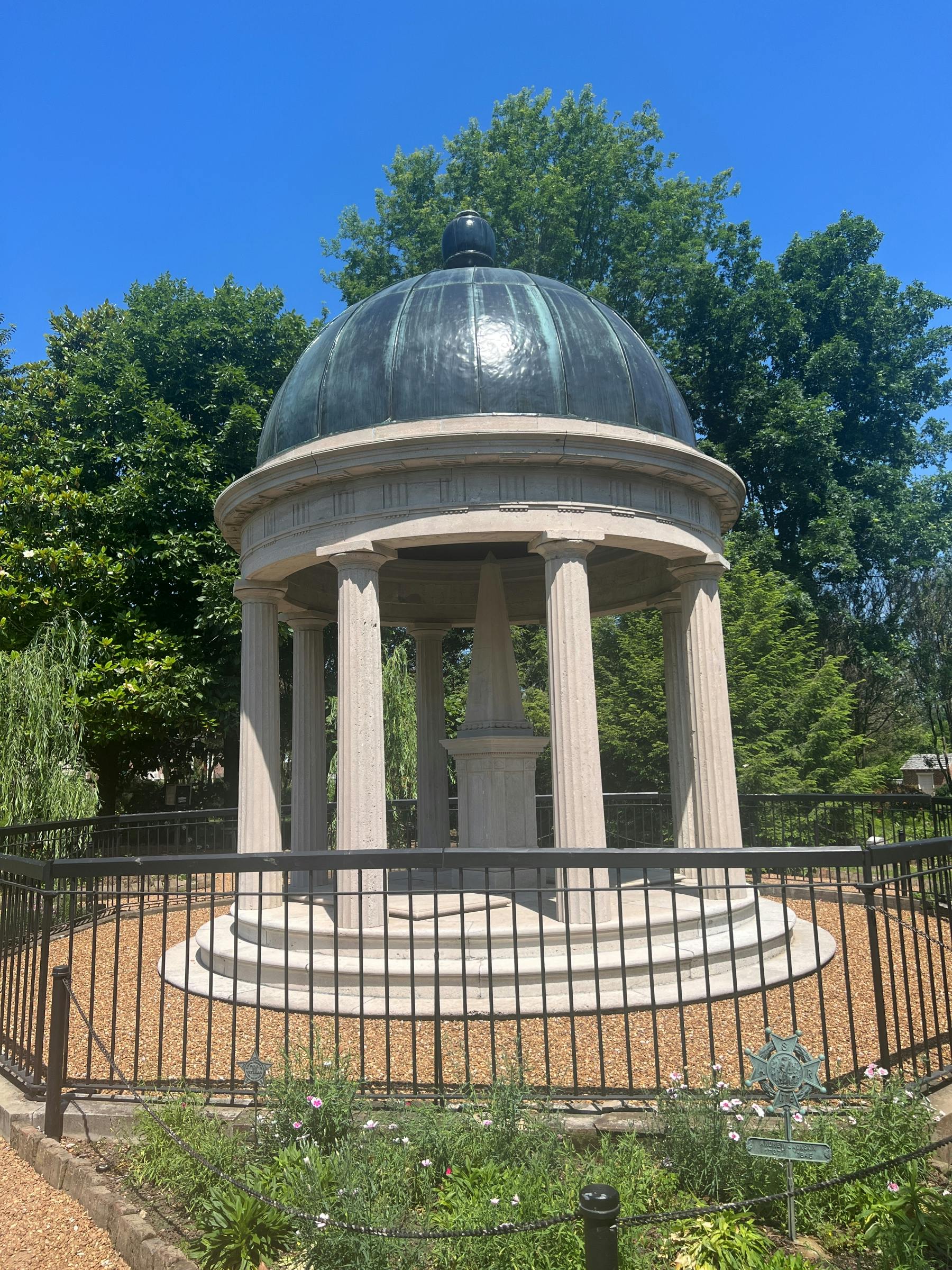 A domed gazebo with columns, surrounded by a black fence, set in a garden with trees and a bright blue sky.