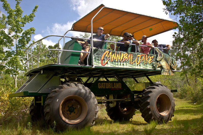 Visitors ride an elevated, large-wheeled vehicle labeled "Cannibal Jake" at Gatorland, under a yellow canopy.
