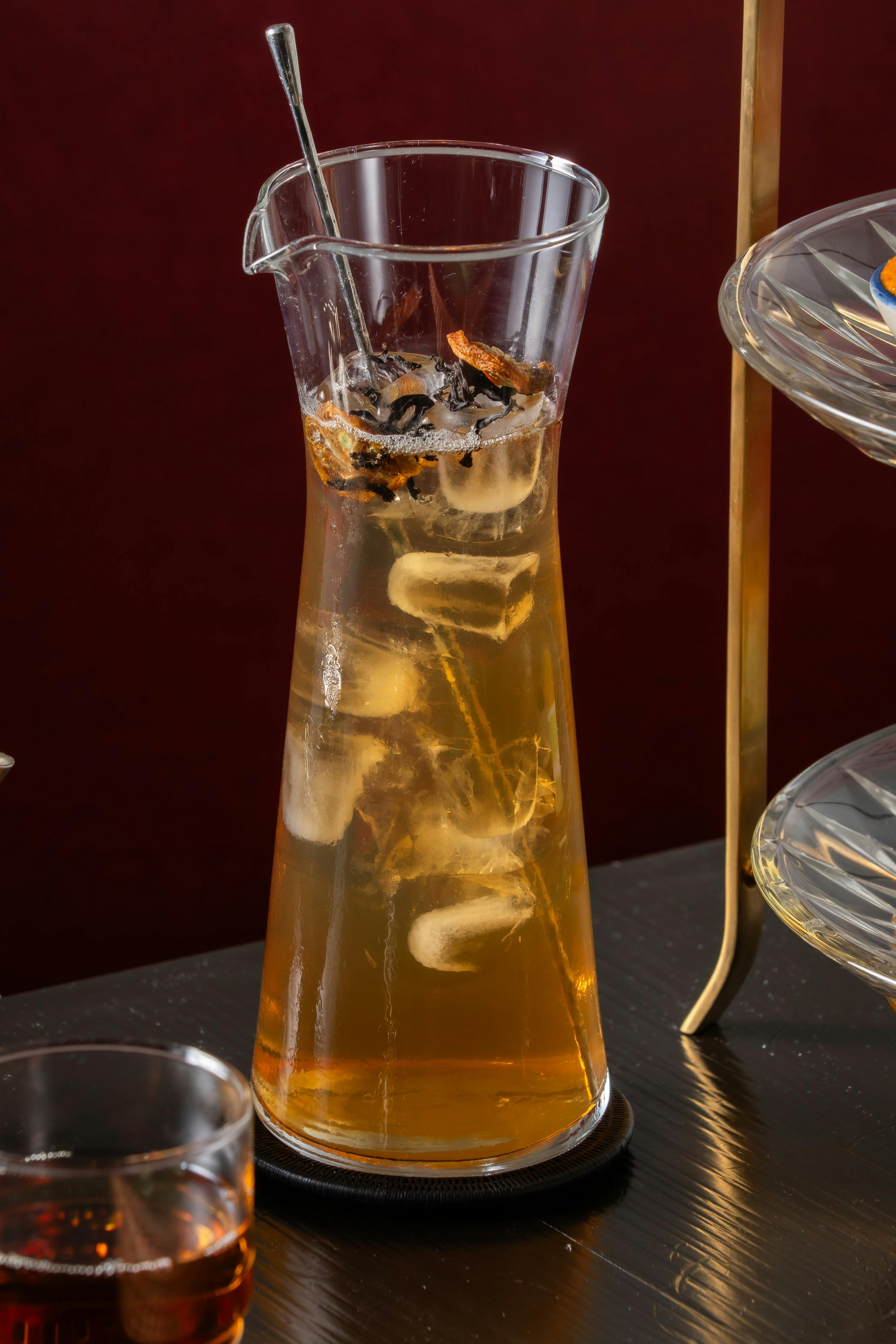 A glass pitcher filled with iced tea, ice cubes, and a garnish, placed on a dark surface near a metal stand.